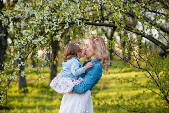 Happy mom and daughter are walking on the street in the garden, in the park, hugging. Mom kisses her daughter. Daughter kisses mom. Green grass, flowering trees in the garden. Mothers Day