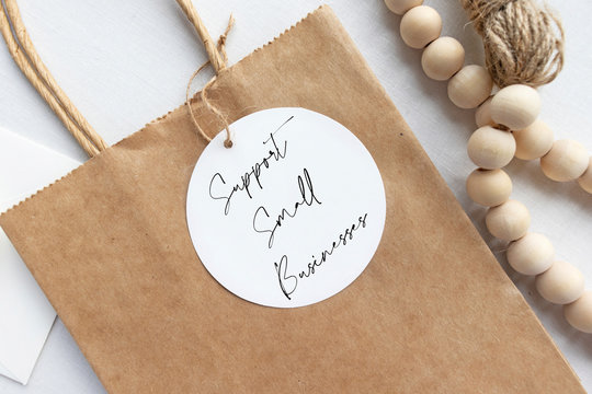 "Support small businesses" text on a gift tag and brown paper bag flat lay with craft props - retail shopping message concept