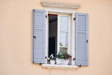 Fototapeta na wymiar Italian window on the pink color wall facade with open wooden grey classic shutters and flowers on the windowsill