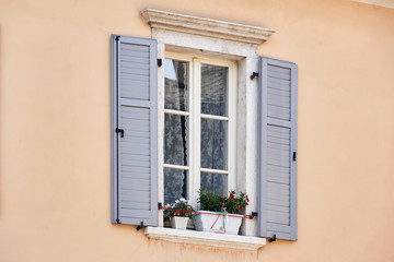 Italian window on the pink color wall facade with open wooden grey classic shutters and flowers on the windowsill