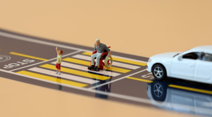 A miniature child with a hand raised and a miniature man  in a wheelchair crossing a crosswalk.
