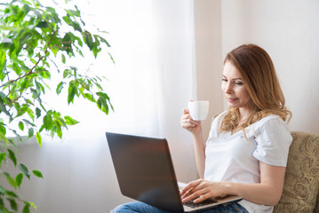 Beautiful young woman working on laptop computer while sitting at the living room, drinking coffee.