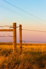 Wooden and Wire Fence posts over a dry grass field with power lines overhead and a rainbow colored sky