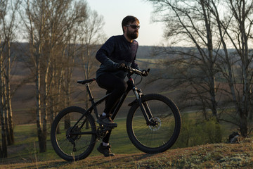 Obraz premium Cyclist in shorts and jersey on a modern carbon hardtail bike with an air suspension fork standing on a cliff against the background of fresh green spring forest