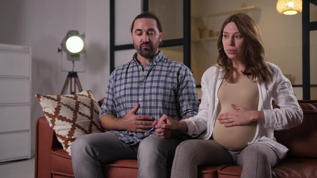 Close-up of focused future parents preparing for childbirth and training special breathing technique while sitting on couch in domestic room. Caring husband breathing with pregnant wife at home