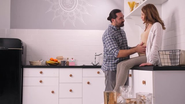 Smiling man caressing big belly, listening to unborn baby and talking to beloved pregnant wife sitting on kitchen counter. Happy couple expecting baby enjoying threesome communication in home kitchen