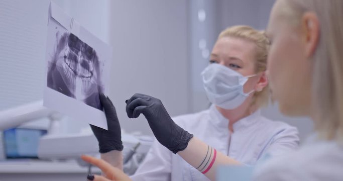 Female dentist shows patient x-ray snapshot of teeth. Dentist says which teeth should be treated and points to them. Modern dentist office.