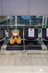 The brown teddy bear wearing a medical face mask sitting on the public chair alone and away from others.