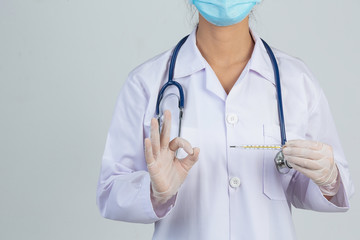 Beautiful young doctor is wearing mask while holding thermometer with rubber gloves on gray background.