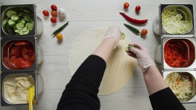 Top view of chef hands in white gloves puts cucumbers on doner kebab shawarma in pita or lavash. Shawarma with chicken cutlet, pappers, cheese and vegetable
