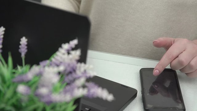 young woman hands with manicure type on black smartphone and laptop with purple flowers on foreground close view