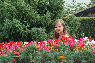 Naklejka premium Little girl with long blond hair in a red dress crouched behind flowers against a background of green bushes and firs and looking away