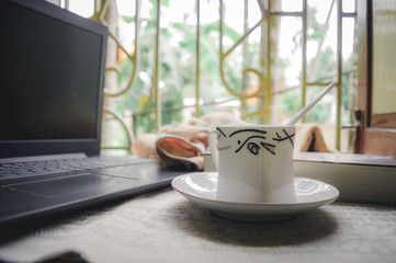 A cup of coffee a book and laptop on window sunlight in morning. A daily routine during social distancing period. Stay home, Work from home, Remote Work and Business Continuity background.