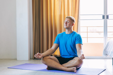Handsome young blond man plays sport at home, a sports guy meditates in a lotus position, holds his hands in mudra in empty room, concept of independent training, fitness and yoga, close-up