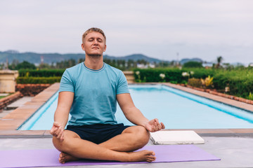 Fototapeta na wymiar Handsome young blond man plays sports outdoors by the pool in tropics during quarantine, guy meditates in a lotus position, holds his hands in mudra, the concept of independent training and fitness