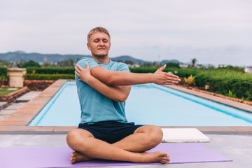 Handsome young blond man plays sports alone outdoors by the pool in tropics during quarantine, guy doing stretching, the concept of independent training, yoga and fitness, close-up