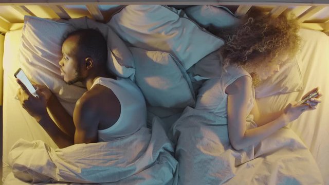 Flat lay of young diverse couple lying back to back to each other in bed and using telephones. Then man switching off the light and putting phone away