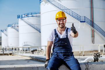 Full length of handsome caucasian workman in overalls and with helmet on head sitting on wall and showing thumbs up. In background are tanks with oil. Refinery exterior.