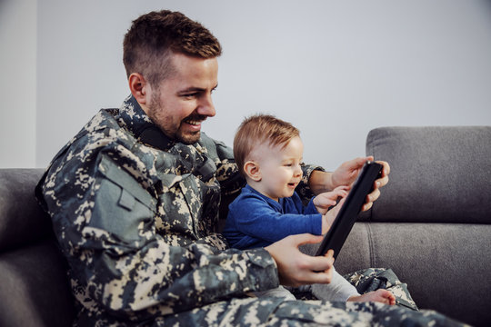 Young soldier making up for the lost time. Man holding son in lap, tablet in hands and plying to his son favorite cartoon. - Powered by Adobe