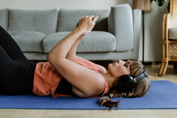 Woman choosing music from her phone while lying on a yoga mat during coronavirus quarantine
