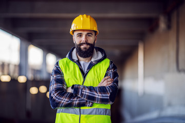 Cute Caucasian bearded construction worker with safety helmet on head in vest standing with arms...