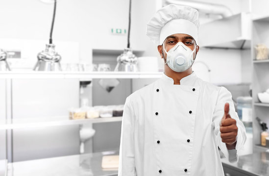 health protection, safety and pandemic concept - indian male chef cook wearing face protective mask or respirator showing thumbs up over restaurant kitchen background