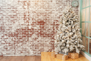 Snow-covered Christmas tree on background of a brick wall with garlands. White fake Christmas tree...