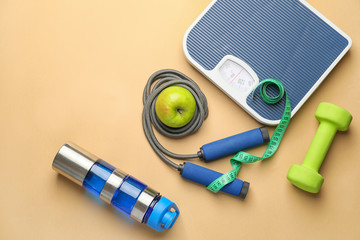 Weight scales with measuring tape, dumbbell, bottle of water, jumping rope and apple on color background