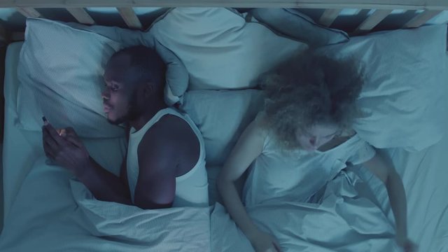 Flat lay African man using mobile phone while his wife is sleeping nearby in bed. Then young woman waking up, taking his phone and they starting a quarrel