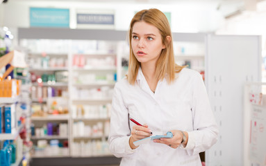 Diligent friendly smiling female pharmacist noting assortment of drugs in pharmacy