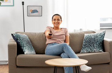 Obraz premium people, race, ethnicity and portrait concept - happy smiling african american young woman with remote control watching tv at home