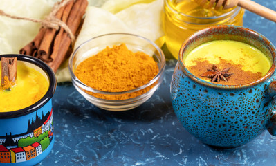 Cups of golden turmeric milk with cinnamon. Ingredients. Yellow napkin and blue backdrop. Close-up, selective focus.