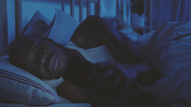 Close up of young African man lying in bed and texting using mobile phone while his girlfriend is sleeping on background - Powered by Adobe