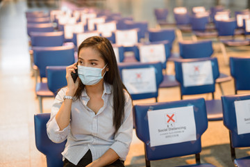 Young Asian tourist woman with mask talking on the phone while sitting with distance at the airport