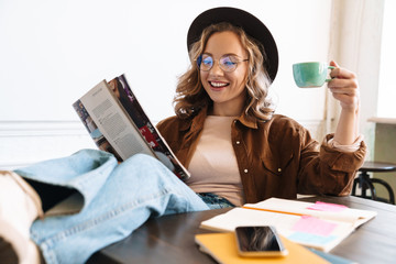 Image of smiling young woman reading magazine while drinking coffee