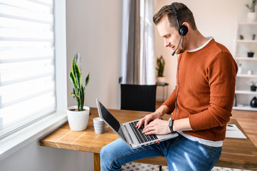 A freelancer guy works from home. Guy with a hands-free headset in the sits on the edge of table, looks at the laptop screen