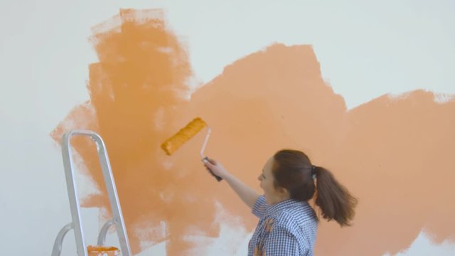 Singleton lifestyle and renovation concept. Young smiling woman with painting roller paints the wall in the apartment and dances