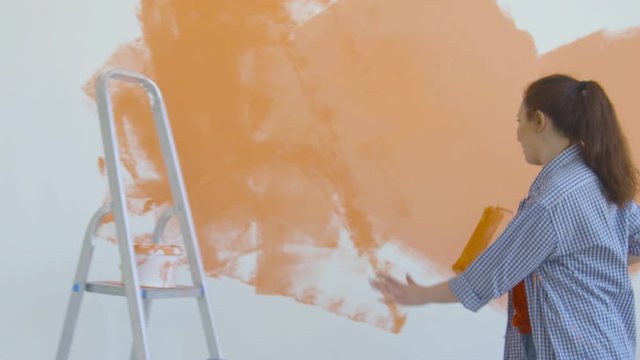 Singleton lifestyle and renovation concept. Young smiling woman with painting roller paints the wall in the apartment and dances