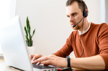 Concentrated guy uses a handsfree headset and laptop for work. He is looking on the screen and...