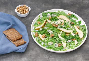 Vegetarian salad with arugula leaves, pear, blue cheese, and walnuts. Some dark bread on blue napkin and more walnuts on the table. Healthy lunch or dinner. 