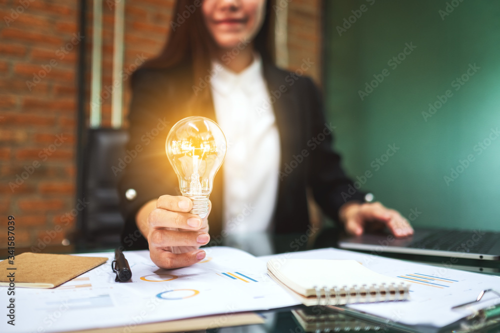 Wall mural closeup image of a businesswoman holding a glowing light bulb while working on laptop computer in of