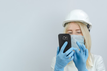 Fototapeta na wymiar young blonde woman in a medical mask and a white construction helmet looks into a black mobile phone close-up, copy space