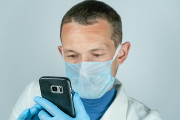 Portrait of a male doctor in a medical gown, mask and gloves holding a black mobile phone and looking into it