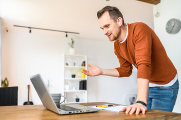 Fototapeta premium Video chat online, video call, zoom. A young bearded guy speaks using a laptop. He is standing, looking at the camera, explaining something and smiling.
