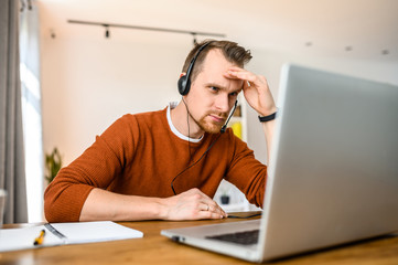 A guy call center operator, support service operator at home office. He uses a headset to talk sits with a cup of coffee at a table, looks at a laptop screen with serious face