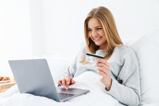 Image of woman holding credit card and using laptop while lying in bed