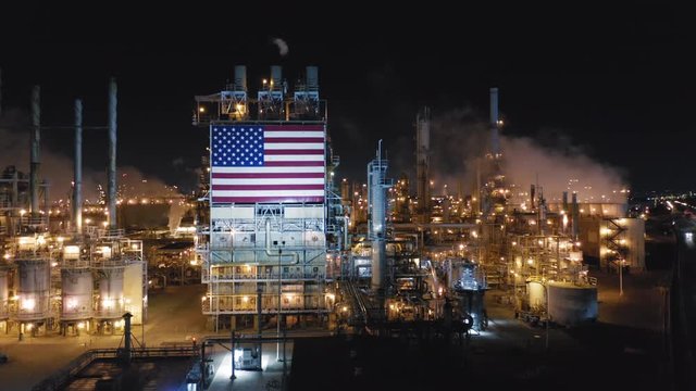 Modern american petroleum refinery at night. Impressive view of huge manufacturing facilities brightly illuminated in the dark. Star-striped american flag is set on one of steel structures. Aerial, 4K