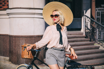 Beautiful blond smiling hipster woman in large wide-brimmed hat riding a retro bicycle in old town...
