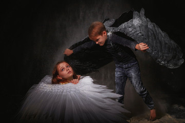 Black and white angel together on a dark background with colored lighting. The concept of war between good and evil. Girl and boy with angel wings during a photo shoot with flour and loose powder