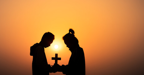 Fototapeta na wymiar Two young christian holding the cross and praying on the mountain with light sunset background, Christians should worship and thank God, christian silhouette concept.